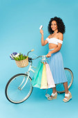 veselý africký americký dívka stojící v blízkosti kola a držící kreditní karty a nákupní tašky na modré, letní koncept 