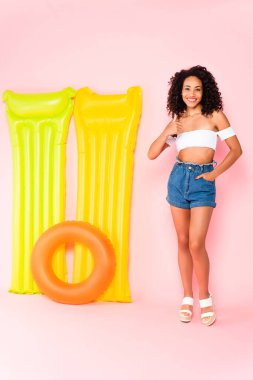 Neşeli Afrikalı Amerikalı kız başparmağını kaldırdı ve cebinde yüzme halkası ve pembe şiltelerin yanında duruyordu. 