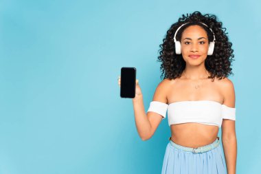 Mutlu Afrikalı Amerikalı kız kablosuz kulaklıkla müzik dinliyor ve elinde mavi ekranda boş ekranlı akıllı telefon tutuyor.