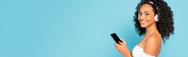 Beyaz ekranlı, kablosuz kulaklıklı neşeli Afrikalı Amerikalı kız ellerinde akıllı telefonuyla mavi ekranda izole edilmiş.