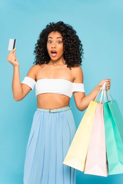 Şok olmuş Afrikalı Amerikalı bir kadın kredi kartı ve alışveriş torbalarını mavi ile tutuyor. 
