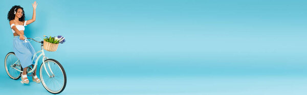 панорамный урожай веселой африканской девушки в наушниках, катающейся на велосипеде и машущей рукой на синей летней концепции 