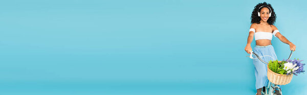 панорамный урожай веселой африканской девушки в наушниках на велосипеде на голубом, летняя концепция 