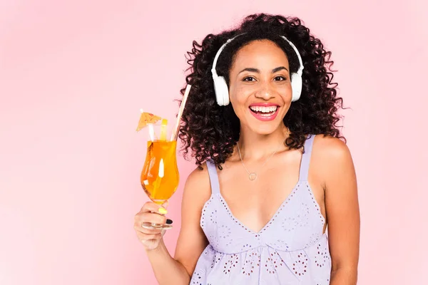 幸せなアフリカ系アメリカ人の女の子でワイヤレスヘッドフォンで音楽を聴きピンクに隔離されたカクテルを保持 — ストック写真