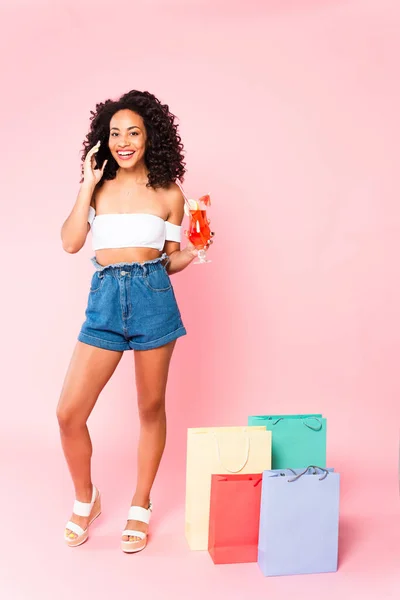快乐的非洲裔美国女人 一边拿着鸡尾酒一边用智能手机说话 一边站在粉色购物袋边 — 图库照片