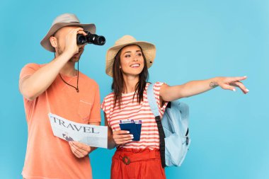 happy girl pointing with finger and holding passports near man with travel newspaper looking through binoculars isolated on blue  clipart