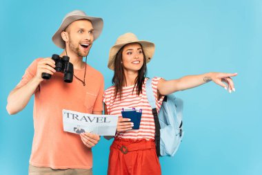 happy girl pointing with finger and holding passports near excited man with travel newspaper and binoculars isolated on blue  clipart