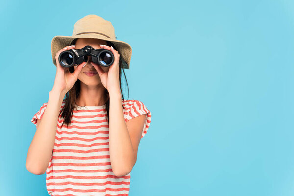 young woman in hat looking through binoculars isolated on blue