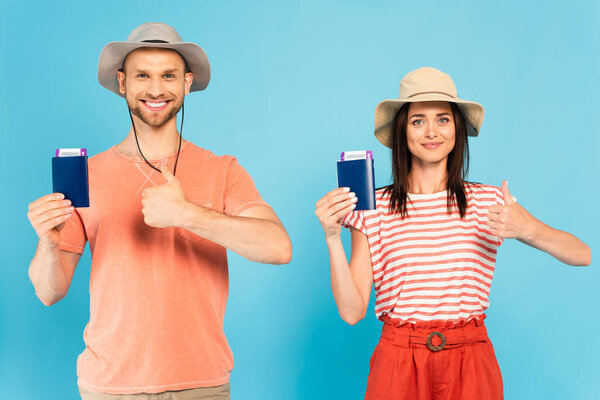 happy man and woman in hats holding passports and showing thumbs up on blue
