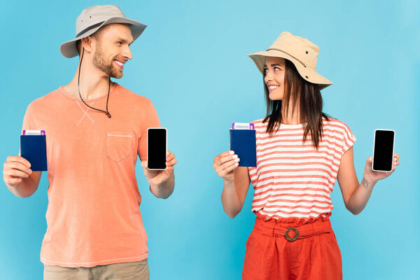 happy couple in hats holding smartphones with blank screen and passports while looking at each other isolated on blue 