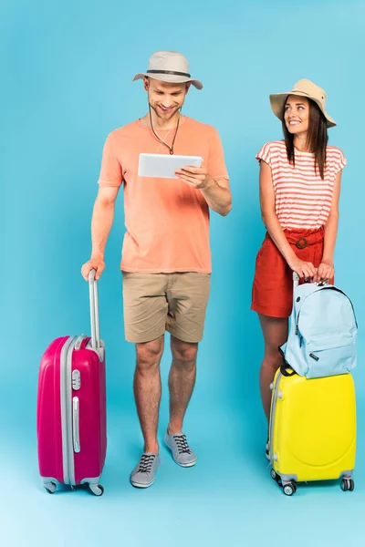 戴帽子的快乐女孩站在蓝色行李旁边看着男人用数码平板电脑 — 图库照片