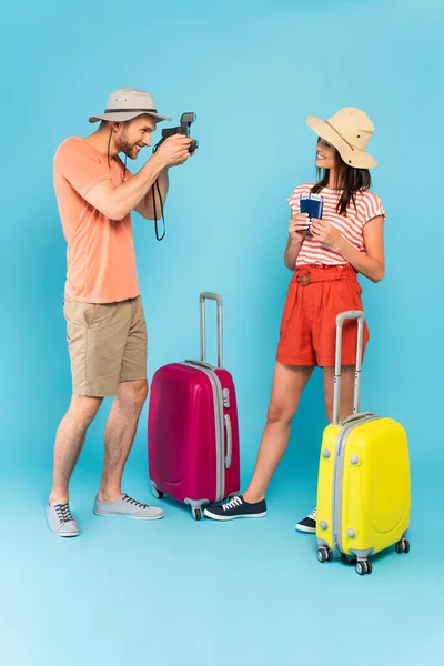 戴帽子的快乐男人拿着老式相机 站在蓝色行李旁边 为拿着护照的女孩拍照 — 图库照片