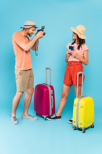 happy man in hat taking photo of girl with passports while holding vintage camera and standing near luggage on blue