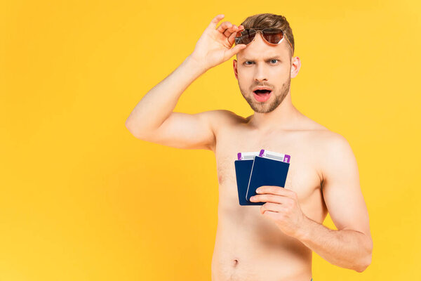 shocked and shirtless man touching sunglasses and holding passports isolated on yellow 