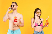 veselý pár v slunečních brýlích a plavkách mluví na smartphonech, zatímco drží koktejly izolované na žluté