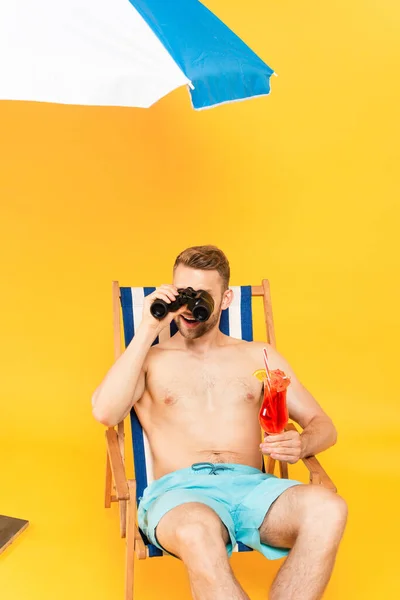 Feliz Hombre Sin Camisa Sosteniendo Cóctel Mirando Través Prismáticos Amarillo — Foto de Stock