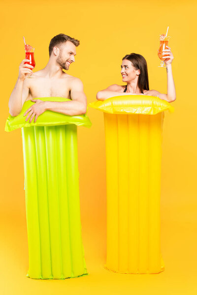 счастливая пара, глядя друг на друга и держа коктейли возле надувных матрасов на желтом
