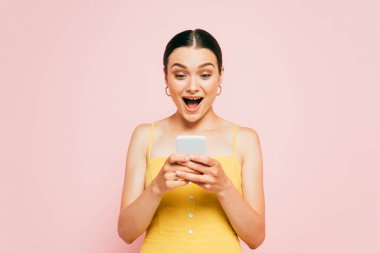 shocked brunette young woman using smartphone isolated on pink clipart
