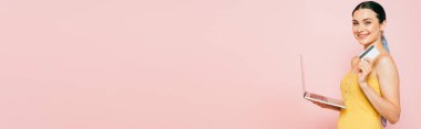 Pembe, panoramik bir karede kredi kartı ve dizüstü bilgisayar tutan esmer genç kadının yan görüntüsü