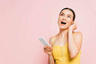 Heyecanlı esmer bir kadın kulaklıkla müzik dinliyor ve elinde pembe bir telefon tutuyor.