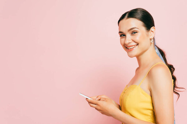 side view of brunette young woman using smartphone isolated on pink