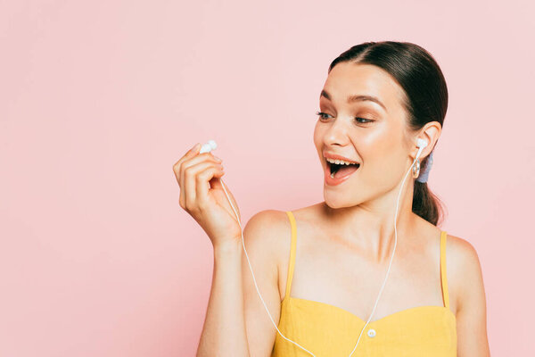 excited brunette young woman looking at earphone isolated on pink