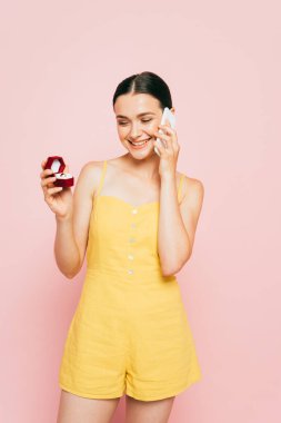 Esmer, genç bir kadın nişan yüzüğüyle kutuyu tutarken pembe bir telefonla konuşuyor.