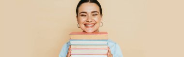 Bej renkli, panoramik bir resimde izole bir şekilde gülümseyen kitaplarla dolu kot gömlekli öğrenci.