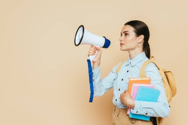 side view of brunette student in denim shirt with books and backpack holding loudspeaker on beige clipart