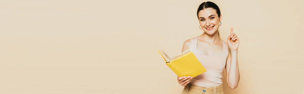 brunette young woman with yellow book showing idea gesture isolated on beige, panoramic shot