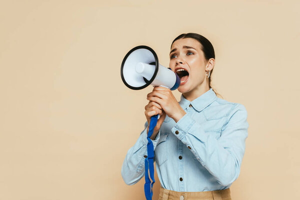 brunette woman in denim shirt screaming in loudspeaker isolated on beige