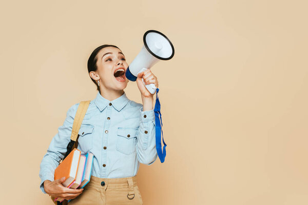 side view of brunette student in denim shirt with books and backpack shouting in loudspeaker on beige