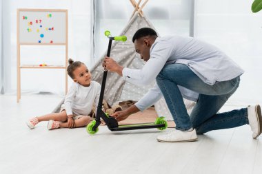 Afrikalı Amerikalı baba, oğlunun yanında scooter tutuyor ve yerde oturuyor.