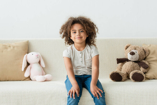curly african american kid looking at camera and sitting on sofa with soft toys