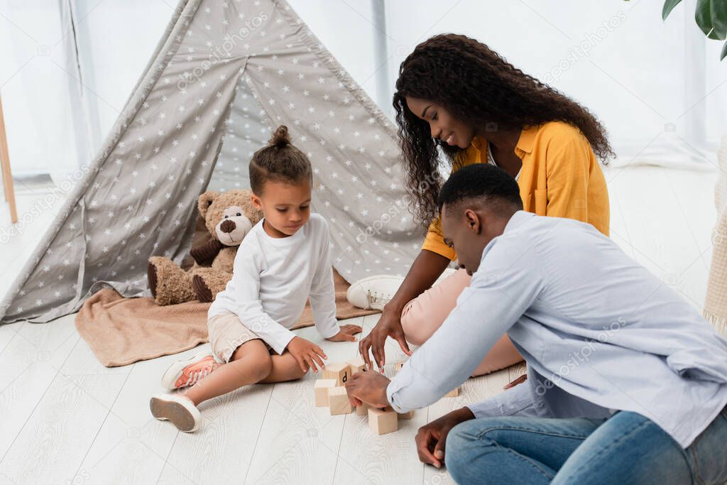 african american parents sitting on floor near son and touching wooden cubes 
