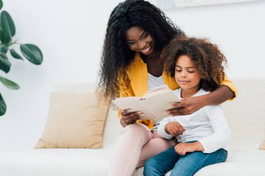Afro-Amerikan annesi kızıyla kanepede otururken kitap okuyor. 