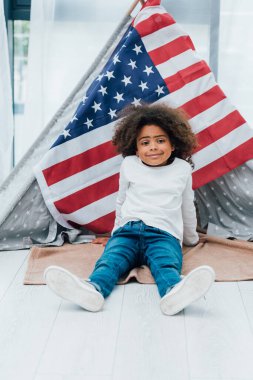 Kıvırcık Afrikalı Amerikalı çocuk Amerika bayrağının yanında yerde oturuyor.