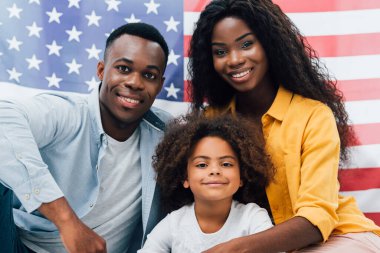 Afro-Amerikan ailesi Amerika bayrağının yanındaki kameraya bakıyor.
