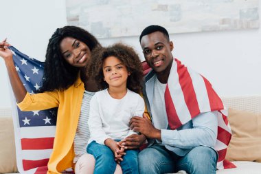 Afro-Amerikan ailesi Amerikan bayrağıyla kaplanmış kanepede otururken kameraya bakıyor. 