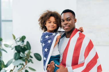 Afro-Amerikan baba ve kızı Amerika bayrağıyla kaplanmış kameraya bakıyorlar. 