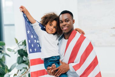 Afrikalı Amerikalı baba ve kız Amerika bayrağına sarılmış kameraya bakıyorlar. 
