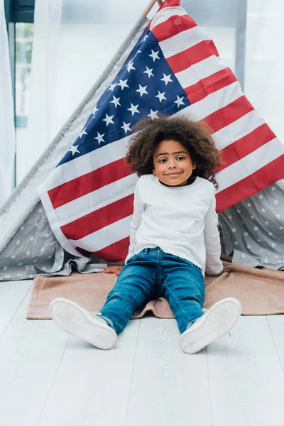 Kıvırcık Afrikalı Amerikalı çocuk Amerika bayrağının yanında yerde oturuyor.
