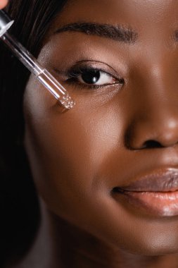 close up view of african american woman applying serum on face isolated on black clipart