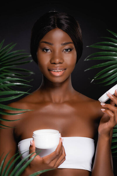 african american woman with cosmetic cream near green palm leaves isolated on black