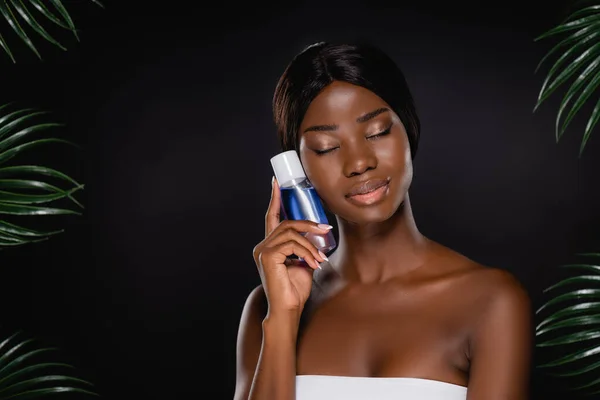 african american woman with closed eyes holding micellar water near green palm leaves isolated on black