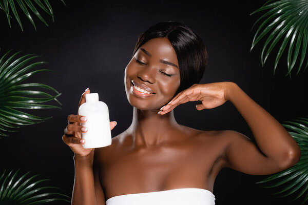Африканская американка держит бутылку лосьона рядом с зелеными пальмовыми листьями, изолированными на черном