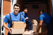 Selective focus of young loader holding cardboard boxes and looking at camera while colleagues unloading truck outdoors 