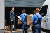 Selective focus of businessman holding clipboard while movers carrying packages near truck and warehouse outdoors