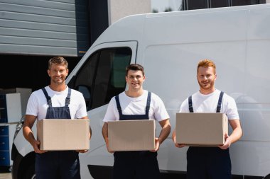Movers in uniform looking at camera and holding cardboard boxes near truck on urban street  clipart