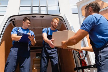 Low angle view of movers holding cardboard box near colleague writing on clipboard  clipart